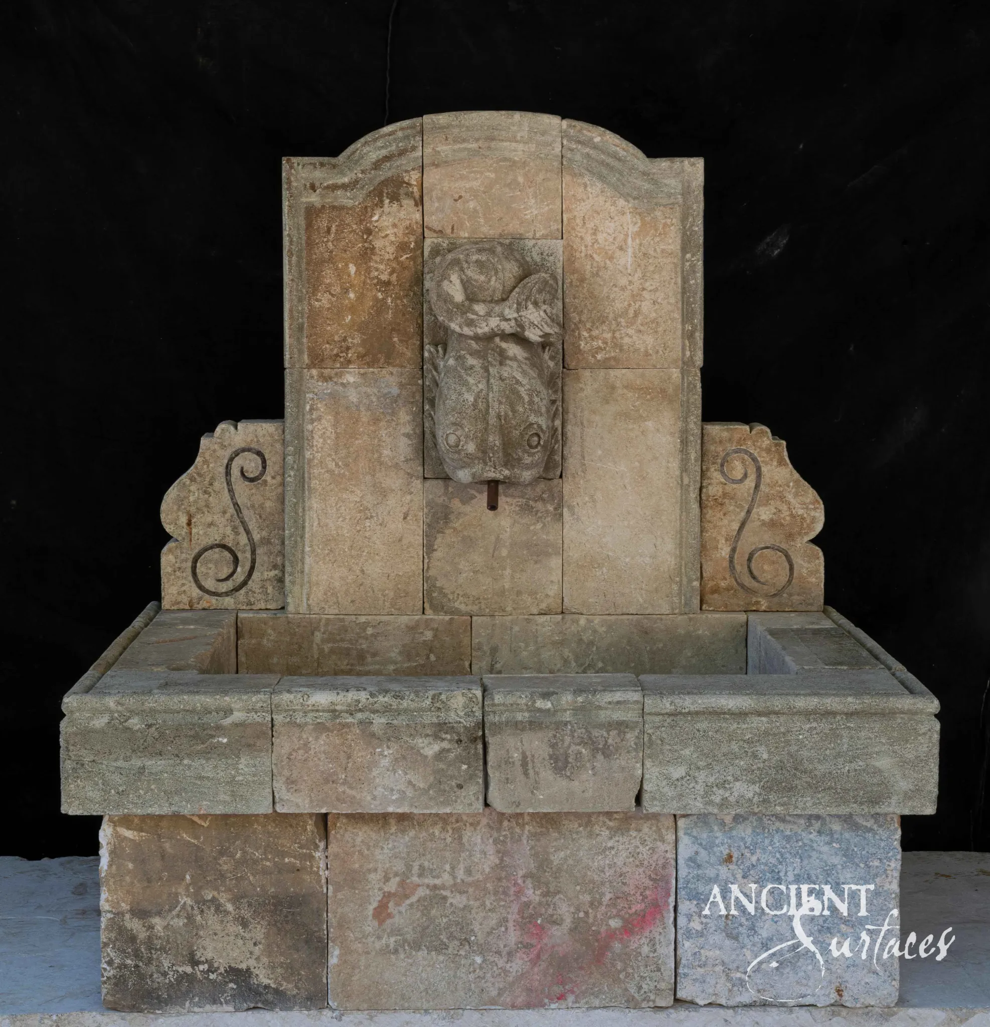 Grey reclaimed limestone wall fountain with a carved dolphin-mask spout and iron scroll wings over a stone trough