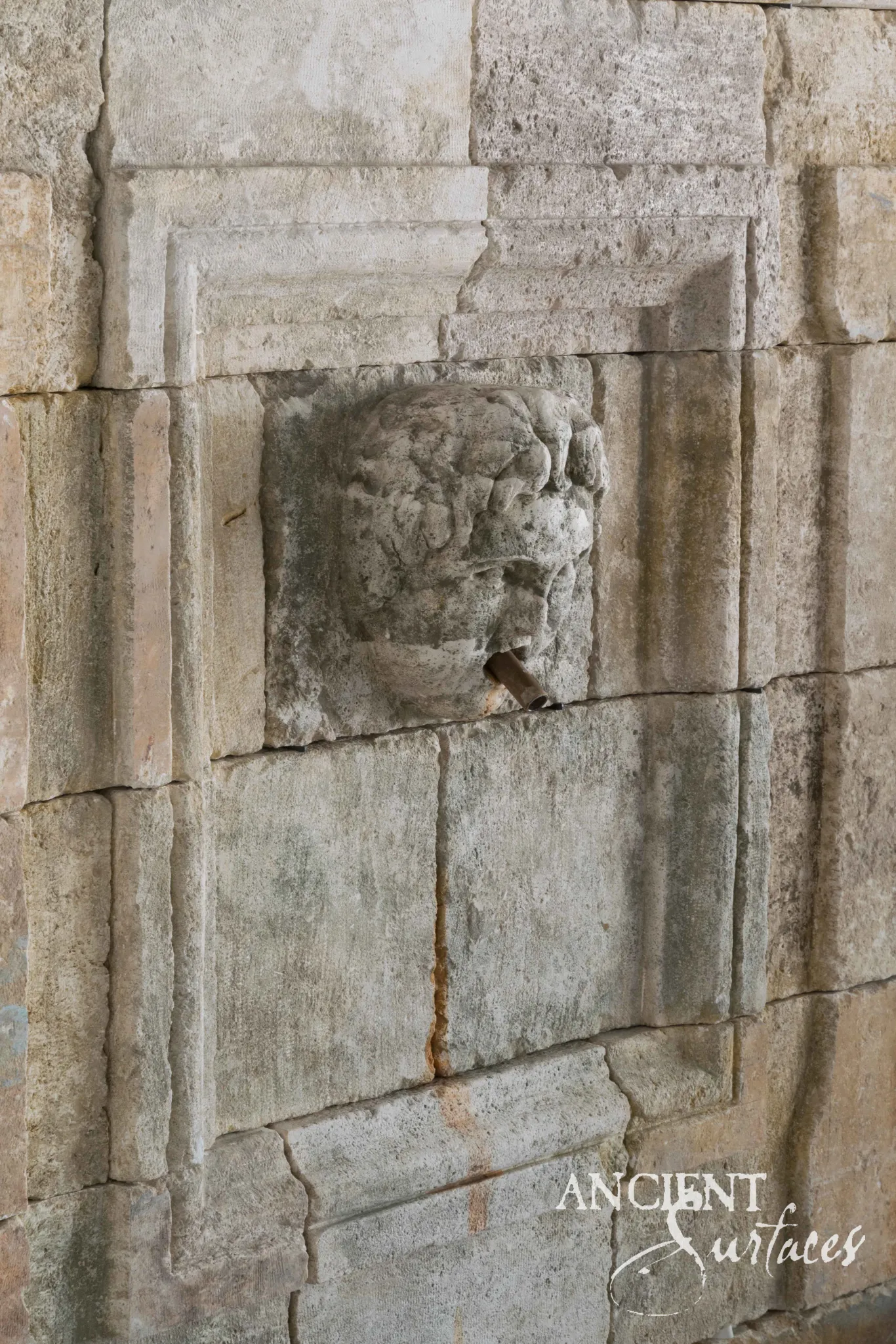 Weathered carved lion-mask spout with an iron pipe set into a reclaimed limestone ashlar wall fountain