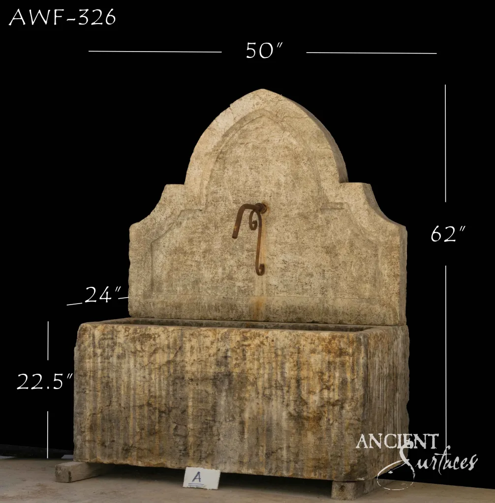 Reclaimed limestone wall fountain with a tall pointed ogee-arch backplate and iron scroll spout over a monolithic trough